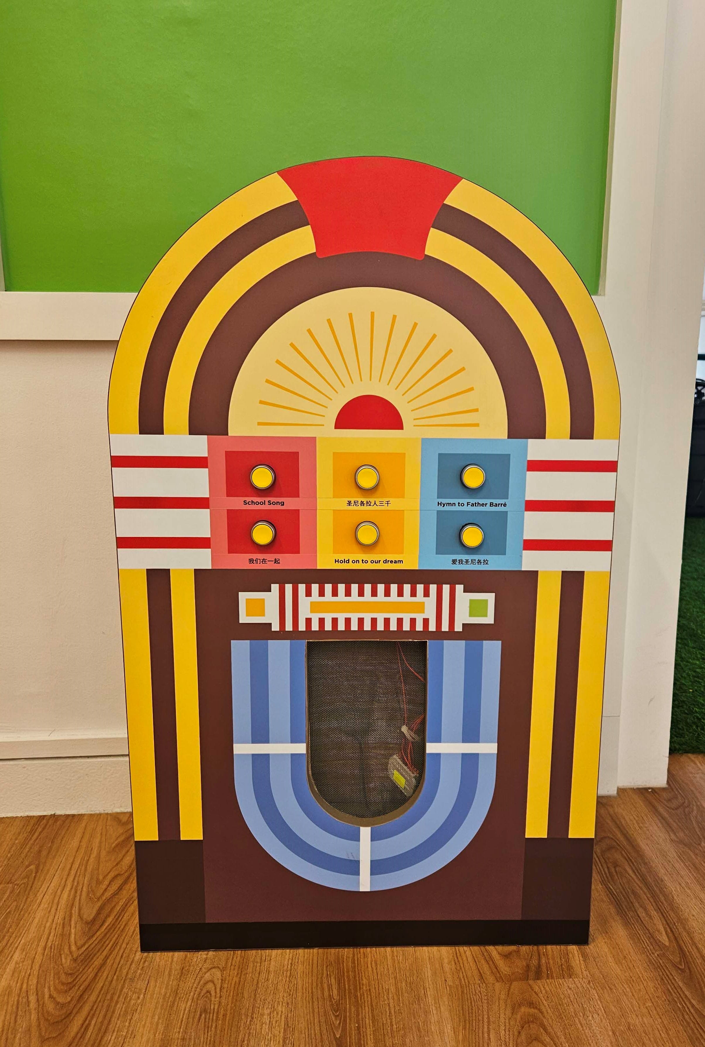A decorative jukebox-shaped interactive exhibit in retro style, featuring six yellow push buttons arranged in three coloured panels — pink, yellow, and blue — each labelled with a school song. 